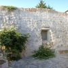 Edificio in pietra con piccola finestra, circondato da piante, illuminato dal sole.